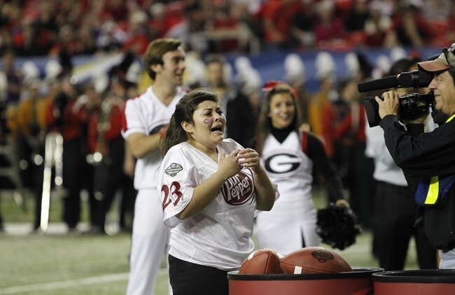 A woman, wearing a white Dr Pepper jersey with the number 23, appears emotional and ecstatic as she stands on a football field during a competition. In front of her, there are footballs and large containers used for a throwing challenge. A cameraman captures her reaction, while cheerleaders and a crowd of spectators in the background cheer her on. The atmosphere is lively and celebratory, suggesting she may have won or completed a successful challenge.