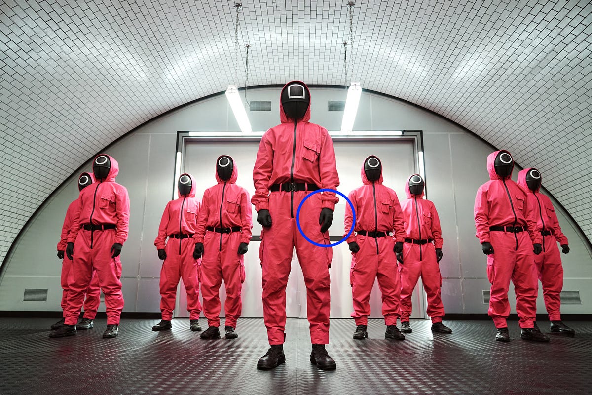 A group of masked figures stands in formation in a stark, brightly lit room with a curved ceiling. Each figure is dressed in a pink hooded jumpsuit and wears black gloves and black shoes, with a mask. The central figure, who appears to be in charge, is marked with a blue circle highlighting a particular detail.