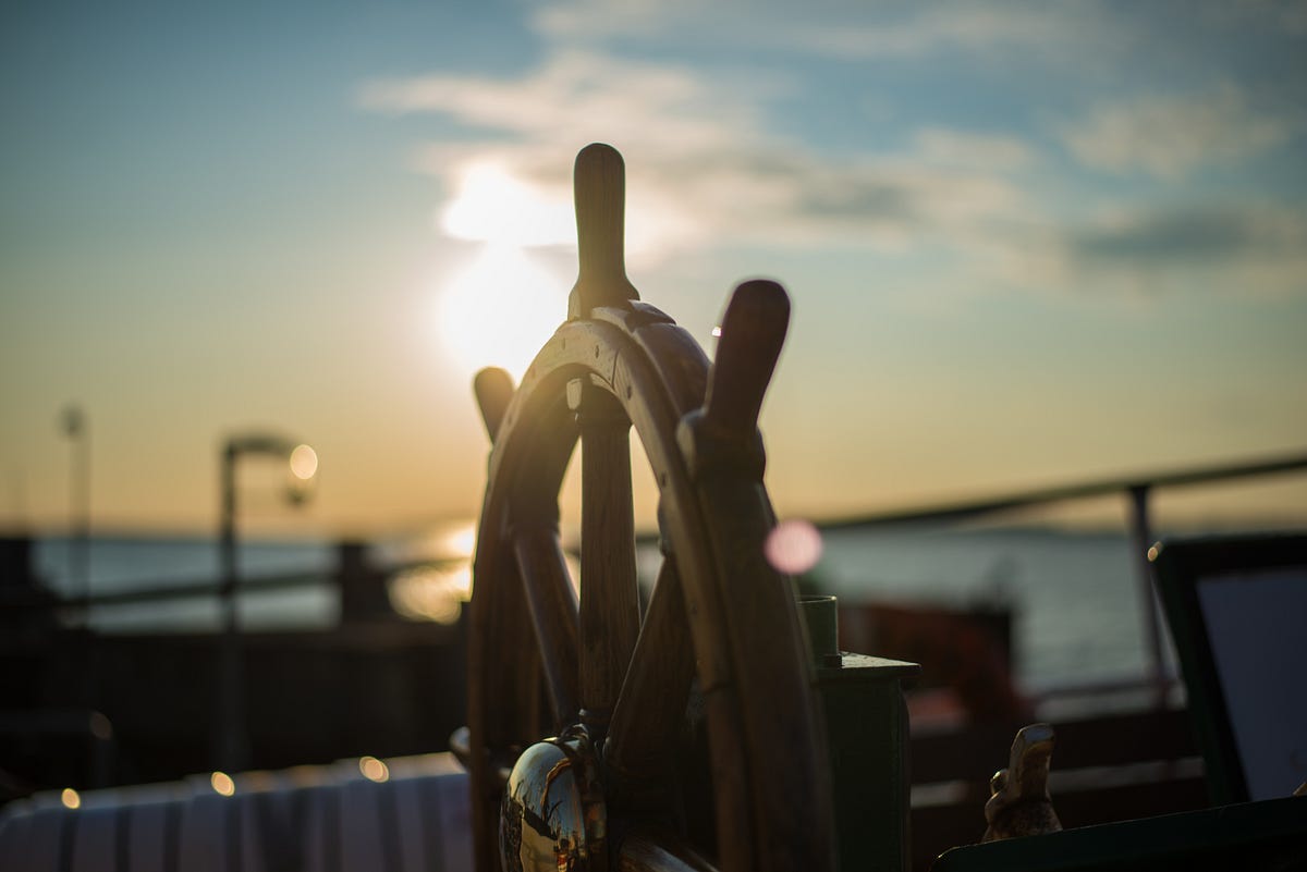 A close-up view of a traditional wooden ship’s wheel, softly lit by the warm rays of the setting sun. In the (bokeh-style) background, the sea stretches out with elements of the dock visible in the immediate vicinity.